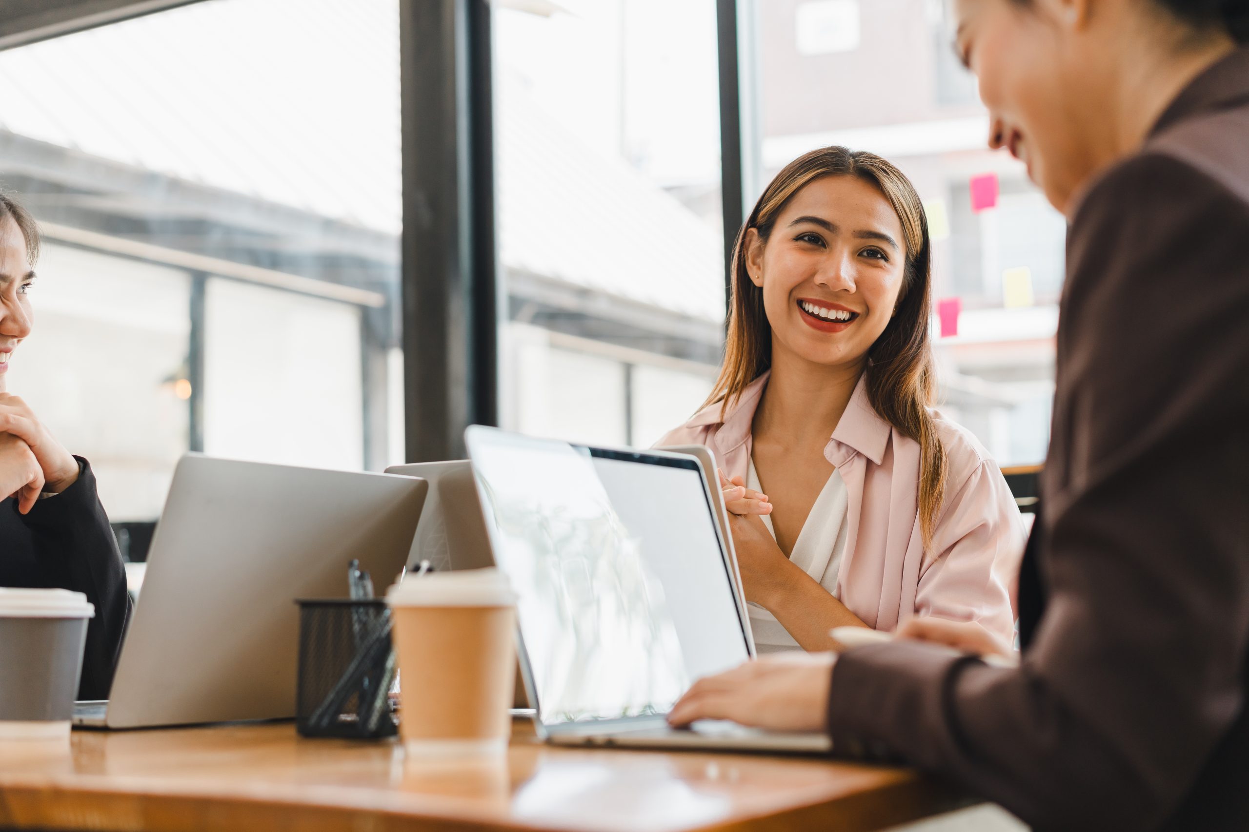 young woman smiles while engaging in conversation with colleagues at modern office setting. atmosphere is collaborative and friendly, with laptops and coffee cups on table.